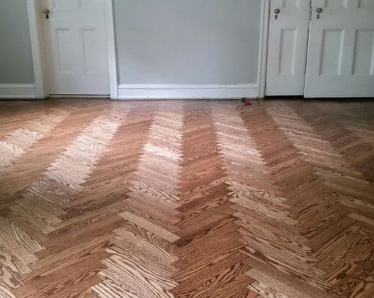Herringbone patterned wooden floor in a minimalist room with gray walls and white doors.