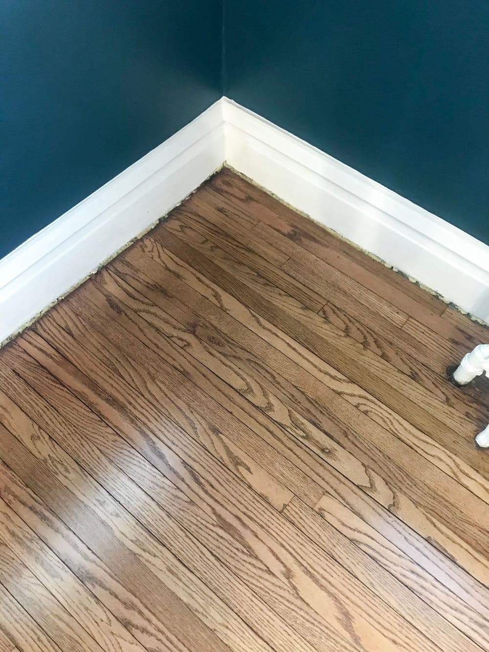 Close-up of hardwood floor corner with baseboard, showcasing wood grain and paint color.