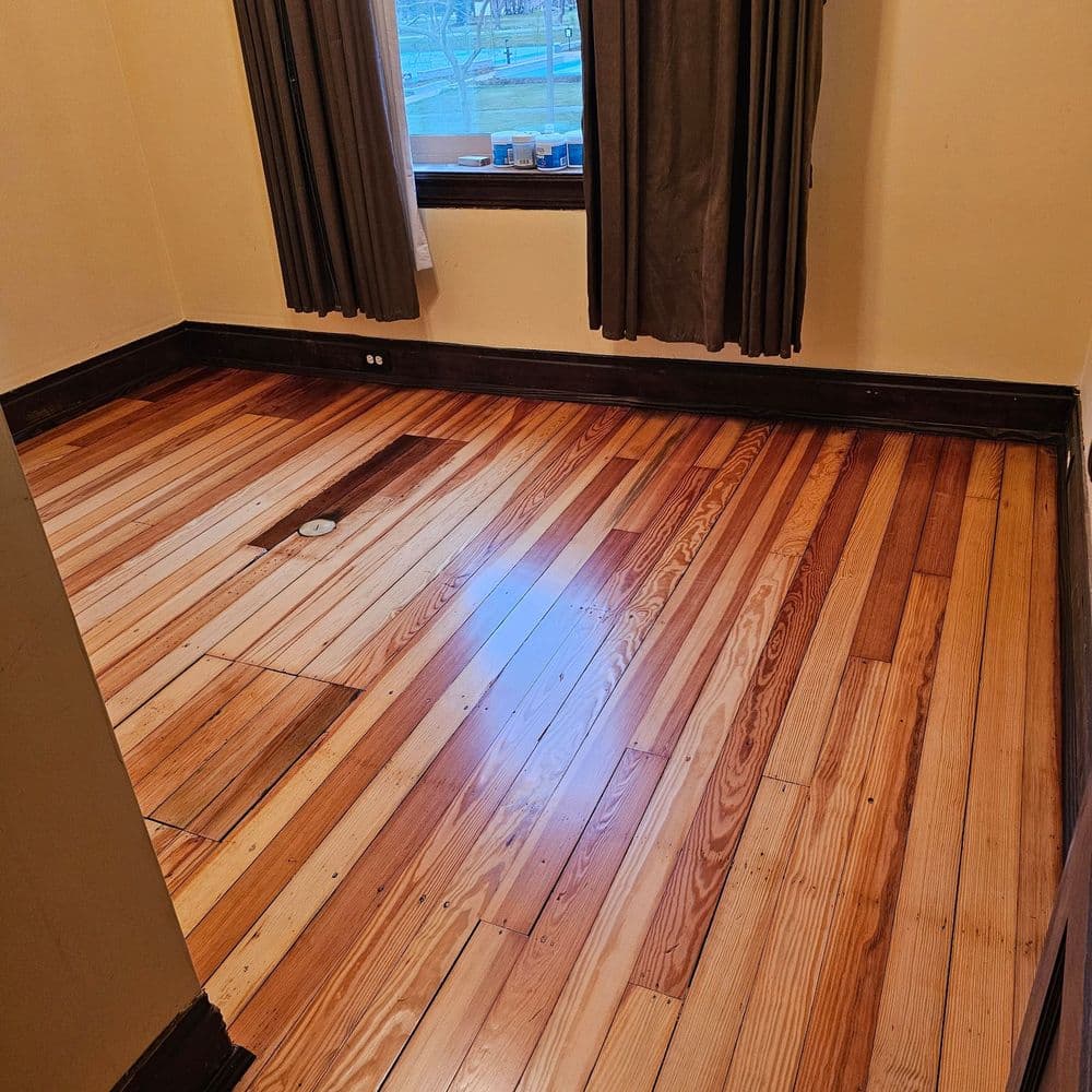 Empty room with hardwood floors and large windows, featuring natural light and beige walls.