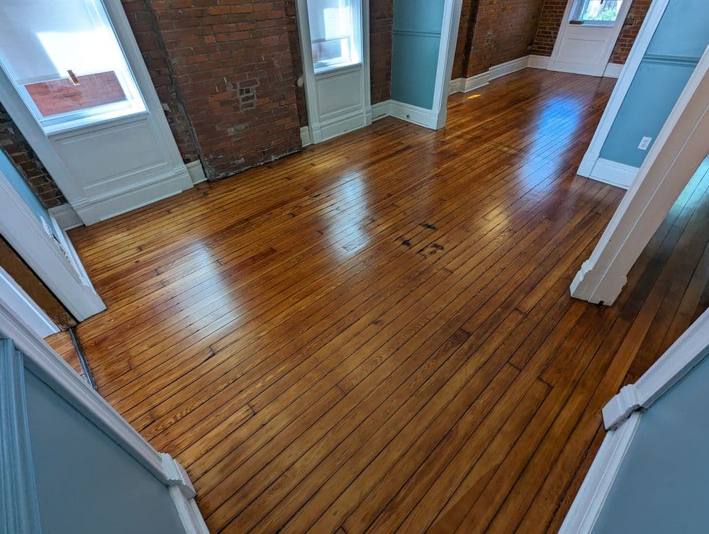 Spacious room with polished hardwood floors and exposed brick walls, featuring large windows.