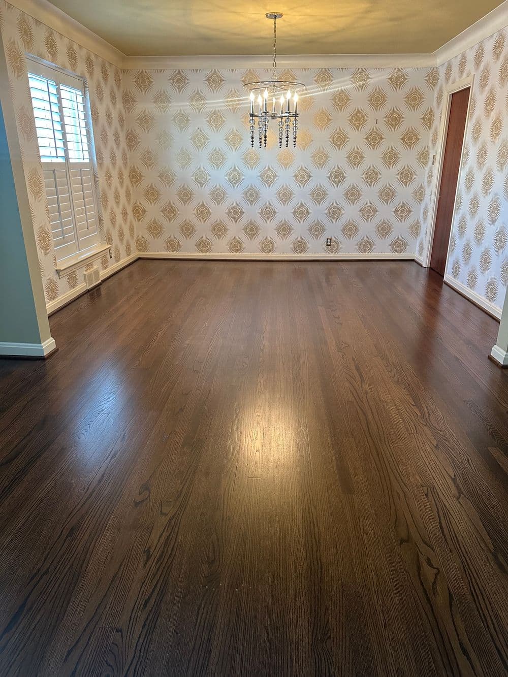 Empty room with dark hardwood flooring, decorative chandelier, and patterned wallpaper.