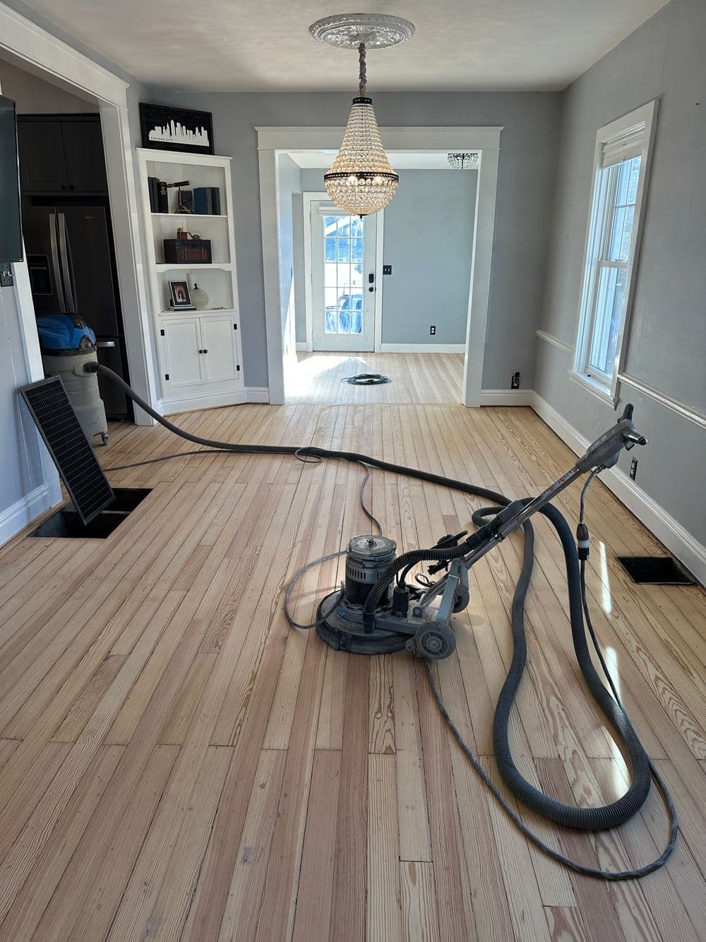 Floor refinishing in a bright room with a sander, showcasing polished wood floors and chandelier.